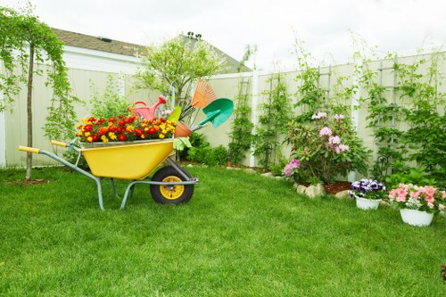 A well-maintained back garden featuring a lush, green lawn with evenly cut grass in the foreground. Against a white panel fence, there is a small flower bed with blooming pink and purple flowers, surrounded by decorative white and pink containers. To the left, a bright yellow wheelbarrow filled with vibrant yellow, red, and orange flowers and gardening tools such as a green watering can and orange trowel are positioned on the grass. Behind the flower bed, a small shrub or young tree with fresh green foliage is visible, adding height to the garden. The background includes a white fence with climbing green plants along its length and a partial view of a neighbouring house roof. The scene is lit with natural daylight, suggestive of a clear or slightly cloudy day, reflecting a tidy outdoor space suitable for gardening and outdoor leisure activities, aligned with professional gardening services in Parsons Green.